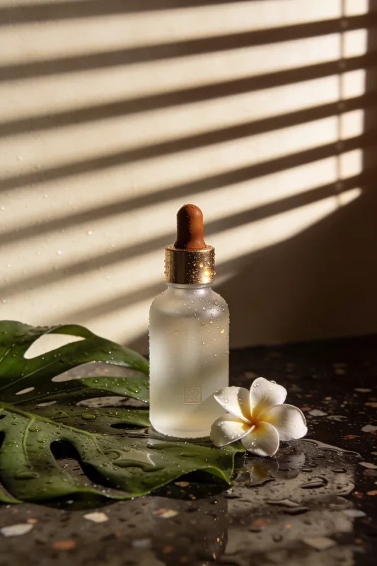 Saga — frosted glass dropper bottle with a brass cap on wet terrazzo, frangipani beside it