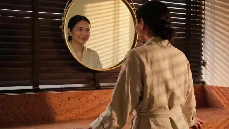 Saga — woman in an oat robe smiling at her reflection in a round brass mirror, warm light through venetian blinds