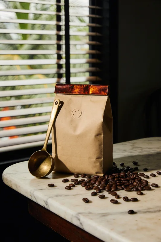 Tanjong Kopi — kraft coffee bag with a blind-embossed mark and terracotta foil band on a kopitiam marble table